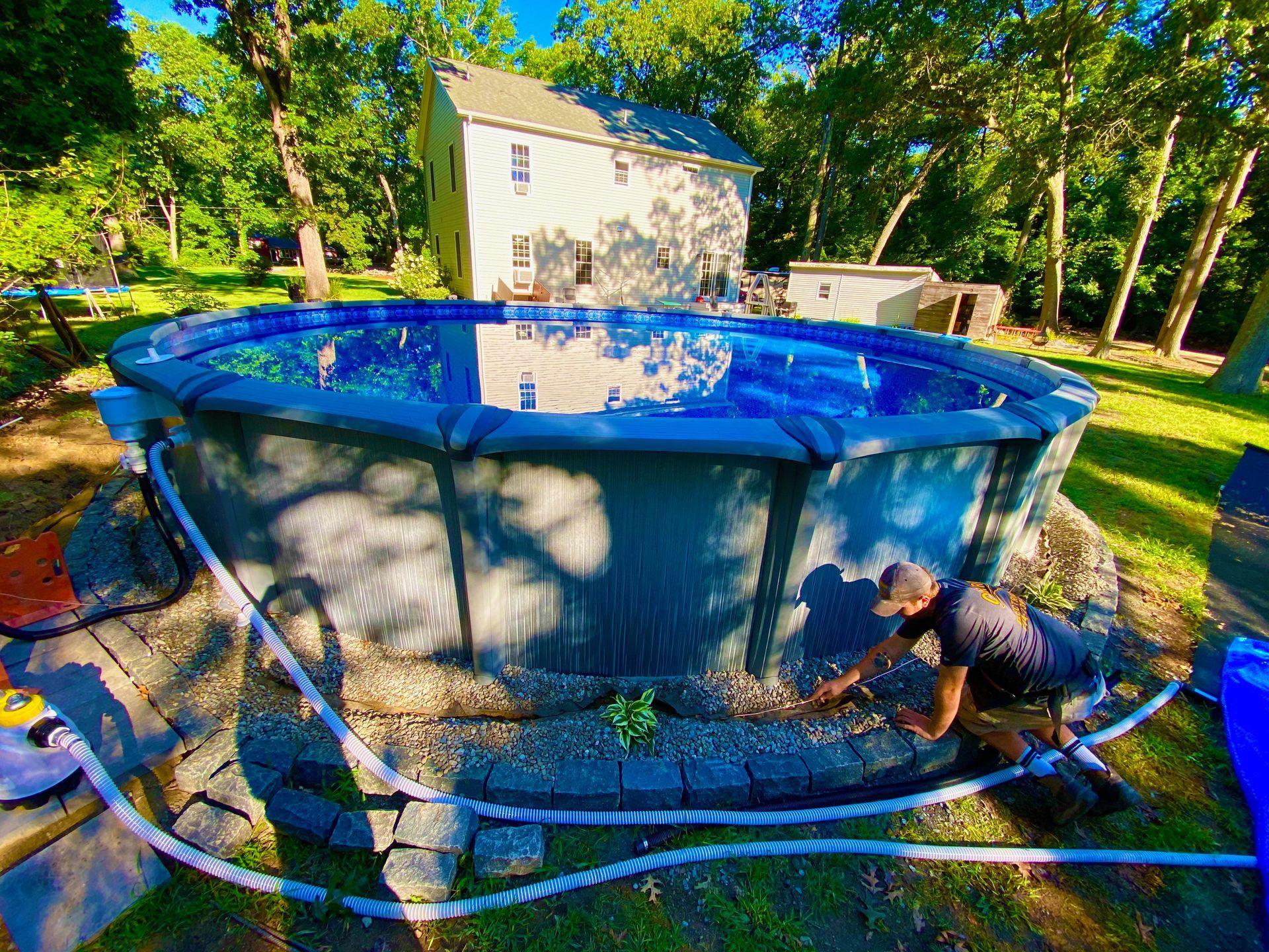 Man working on an above-ground pool surrounded by stone and rope. House and trees in the background.