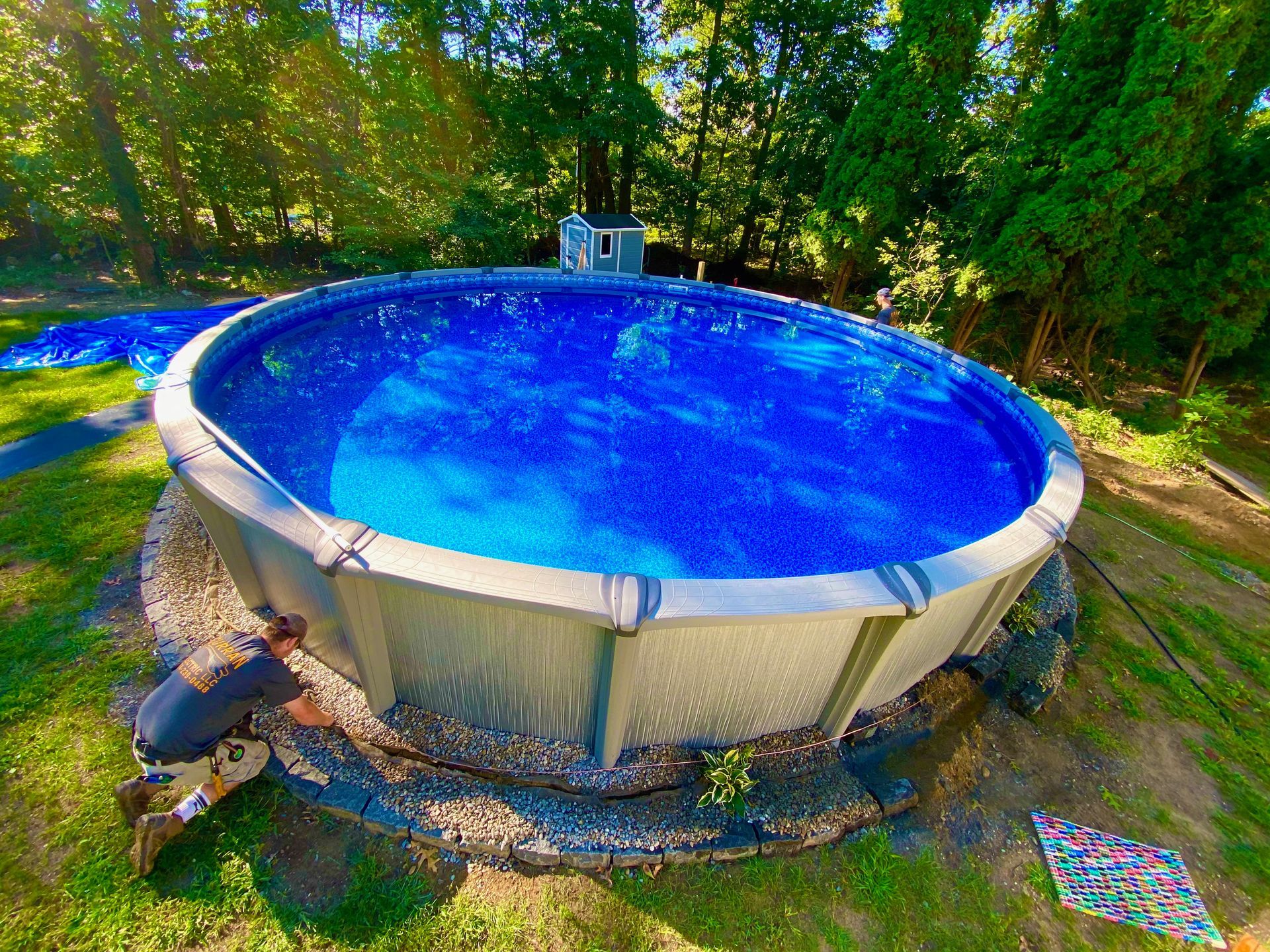 Man installing an above-ground pool.  The round pool is filled with blue water. Surrounding it are rocks and green grass.