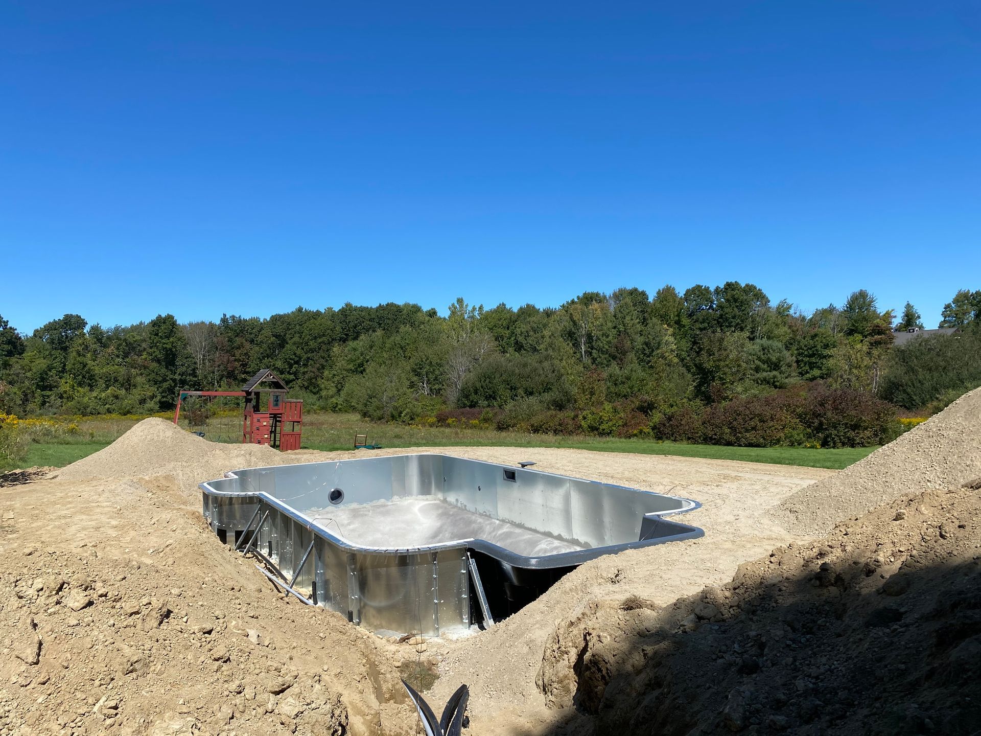 Pool under construction, steel frame in excavated earth. Brown dirt piles surround pool in outdoor setting with trees.