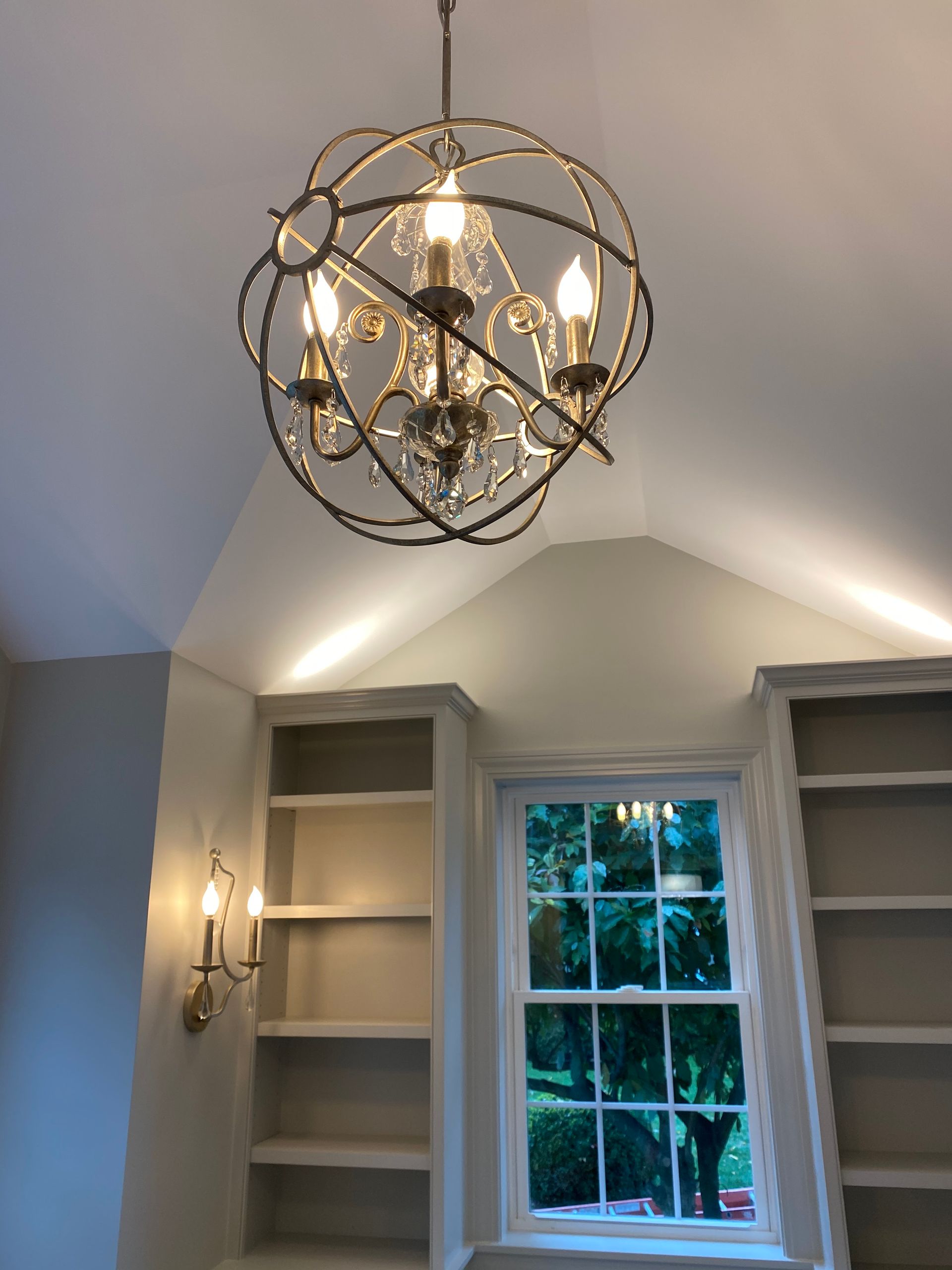Chandelier hangs above a window with built-in bookshelves and sconce. Light-colored walls and ceiling.