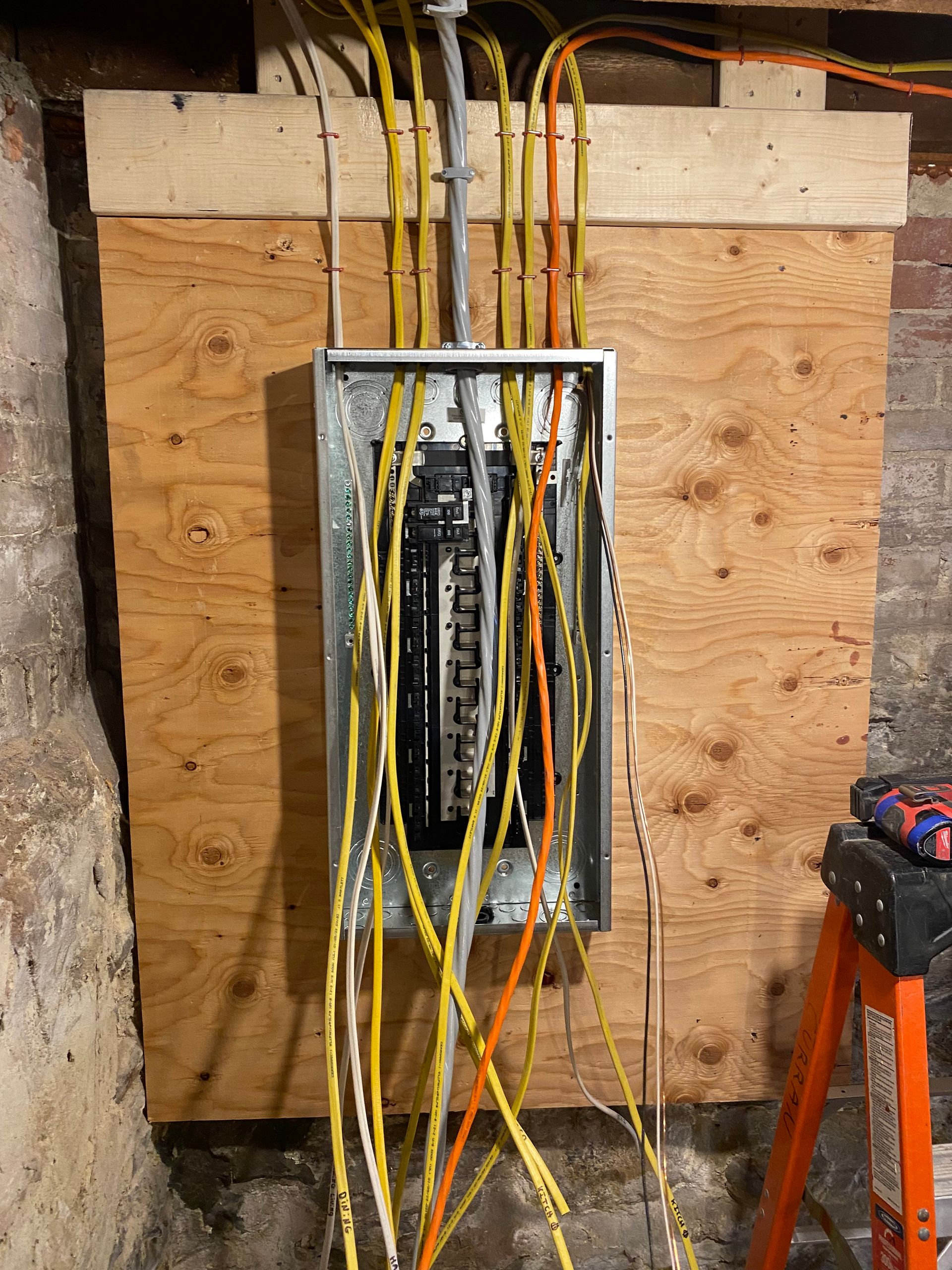 Electrical panel with exposed wires, mounted on plywood against a brick wall.