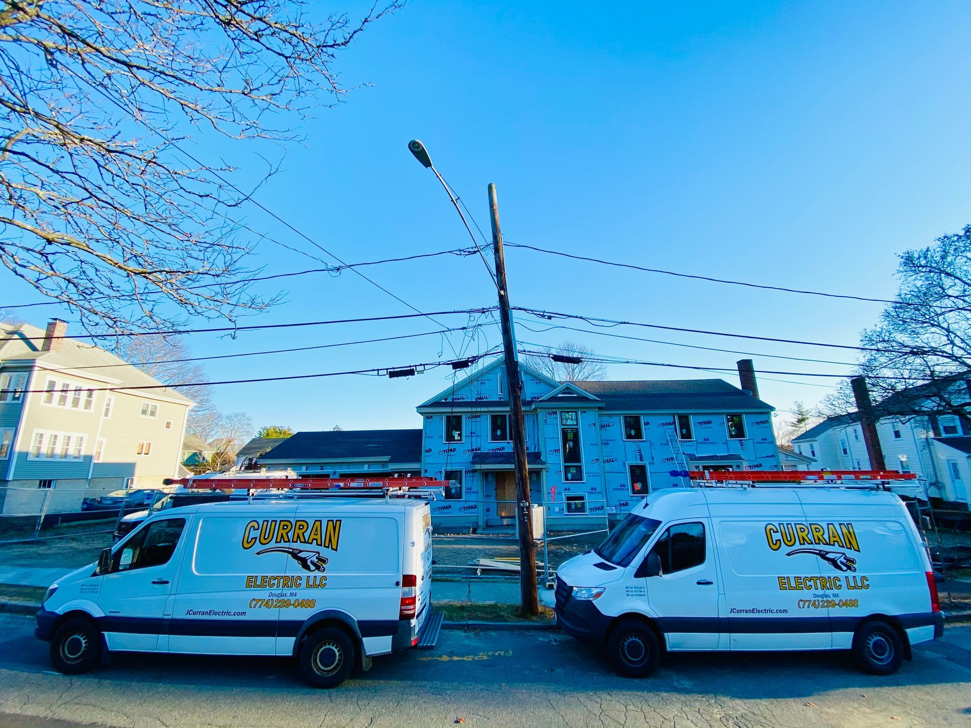 Two white vans with business logos parked on a street; a house and power lines are visible in the background.