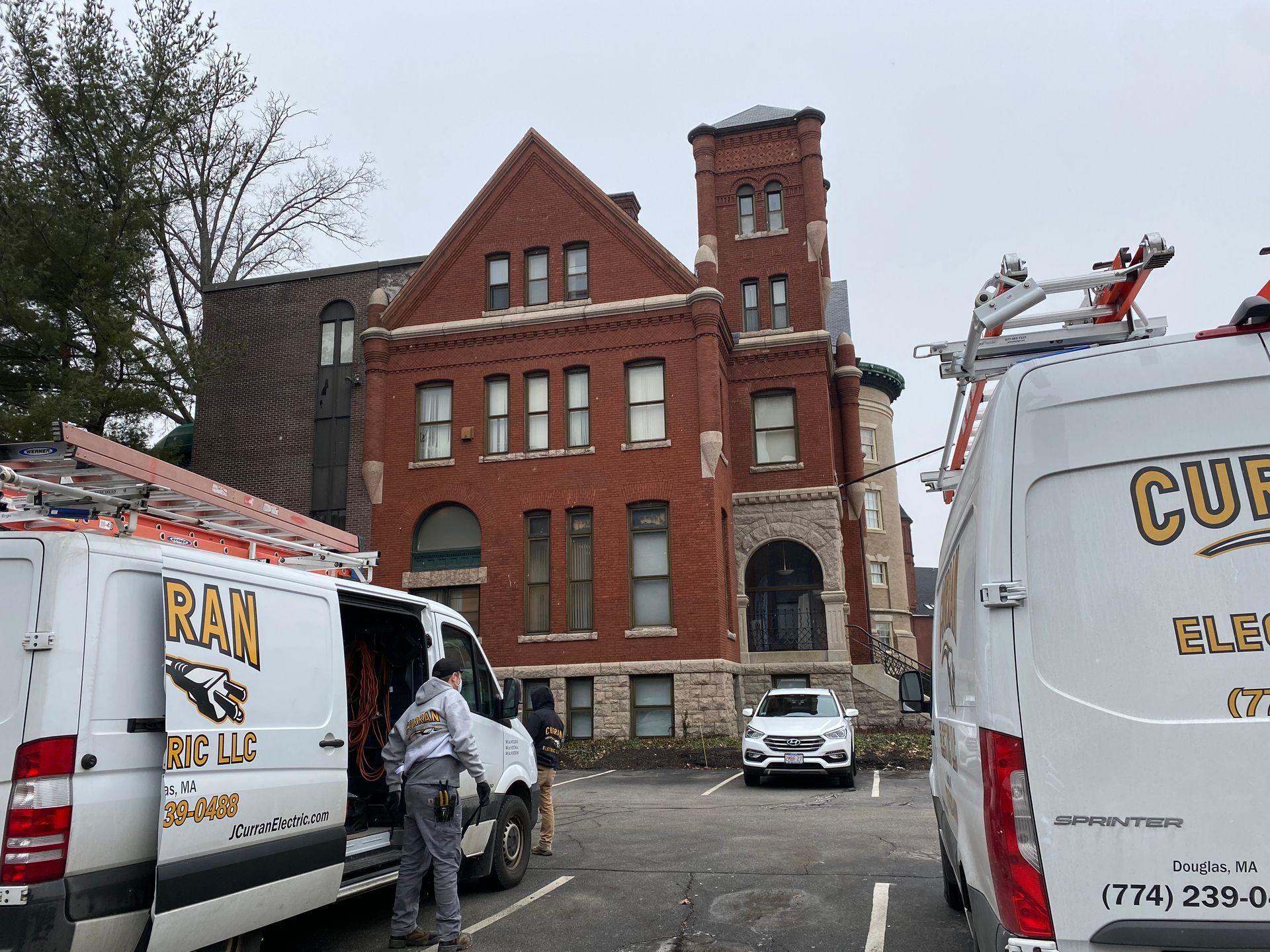 Electrical vans parked near a brick building with a tower. A person stands near a van. Cloudy sky.