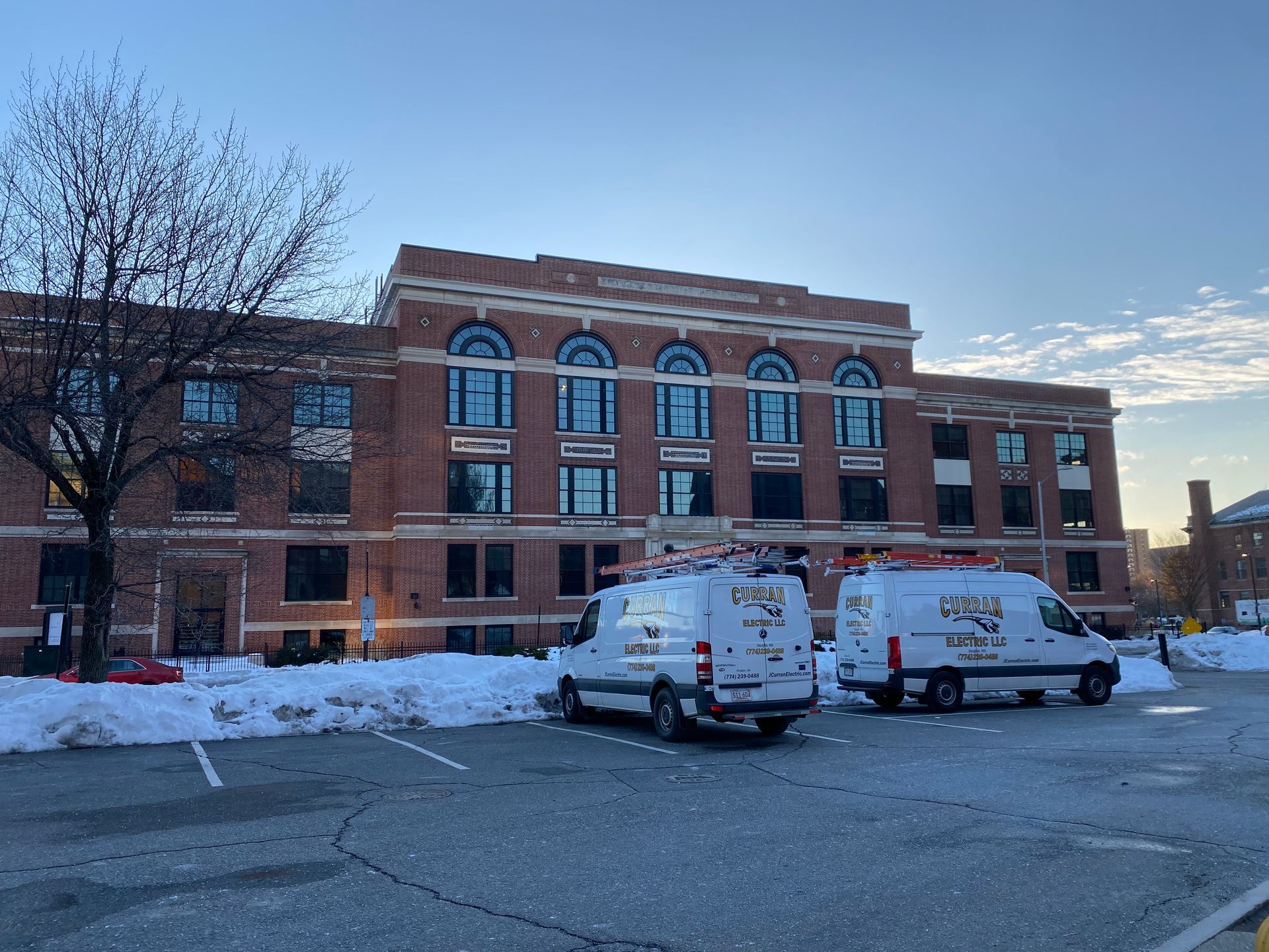 Two white work vans parked in front of a brick building with snow on the ground; blue sky.