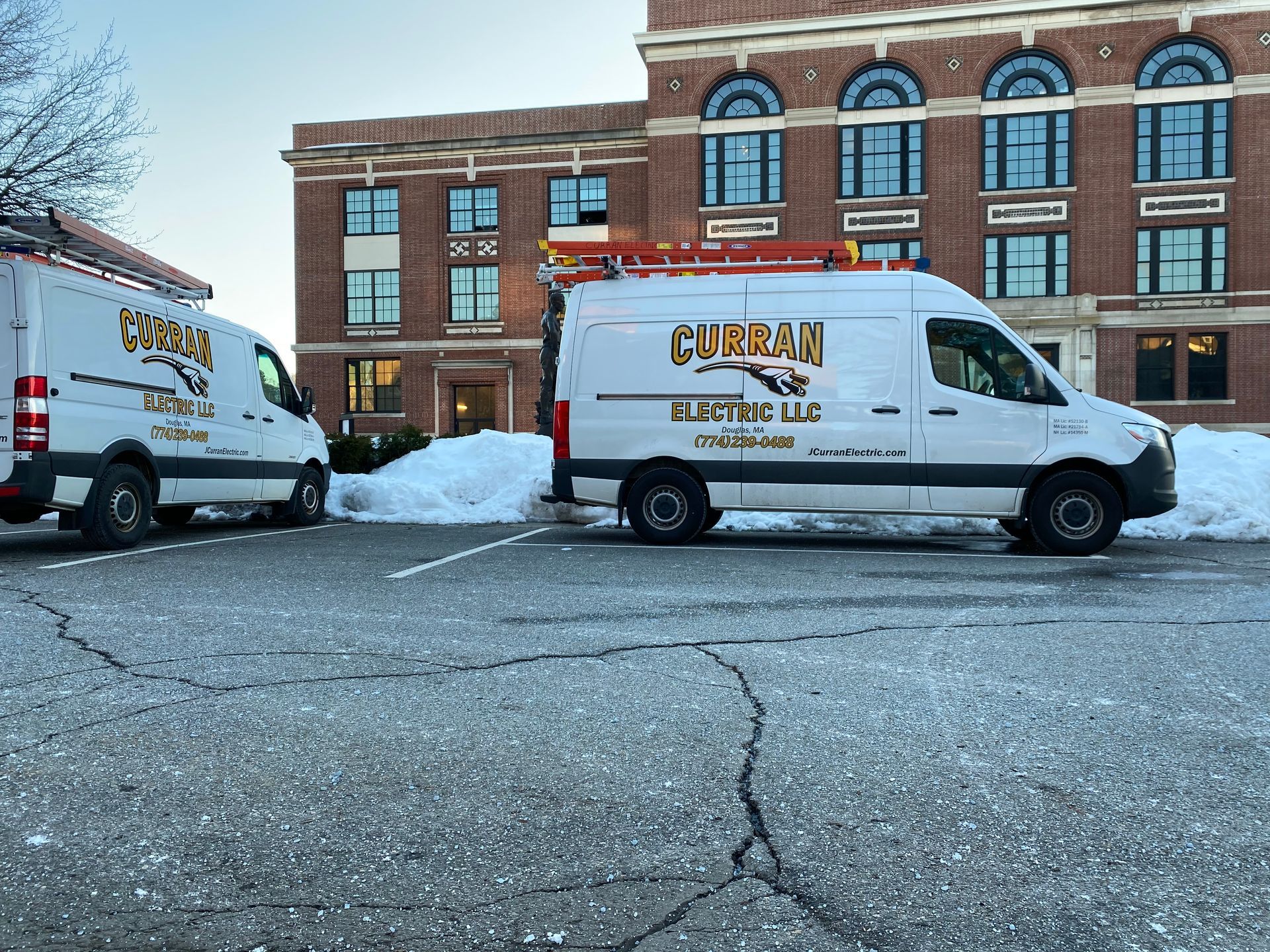 Two white CURRAN ELECTRIC LLC vans parked in front of a brick building with snow on the ground.