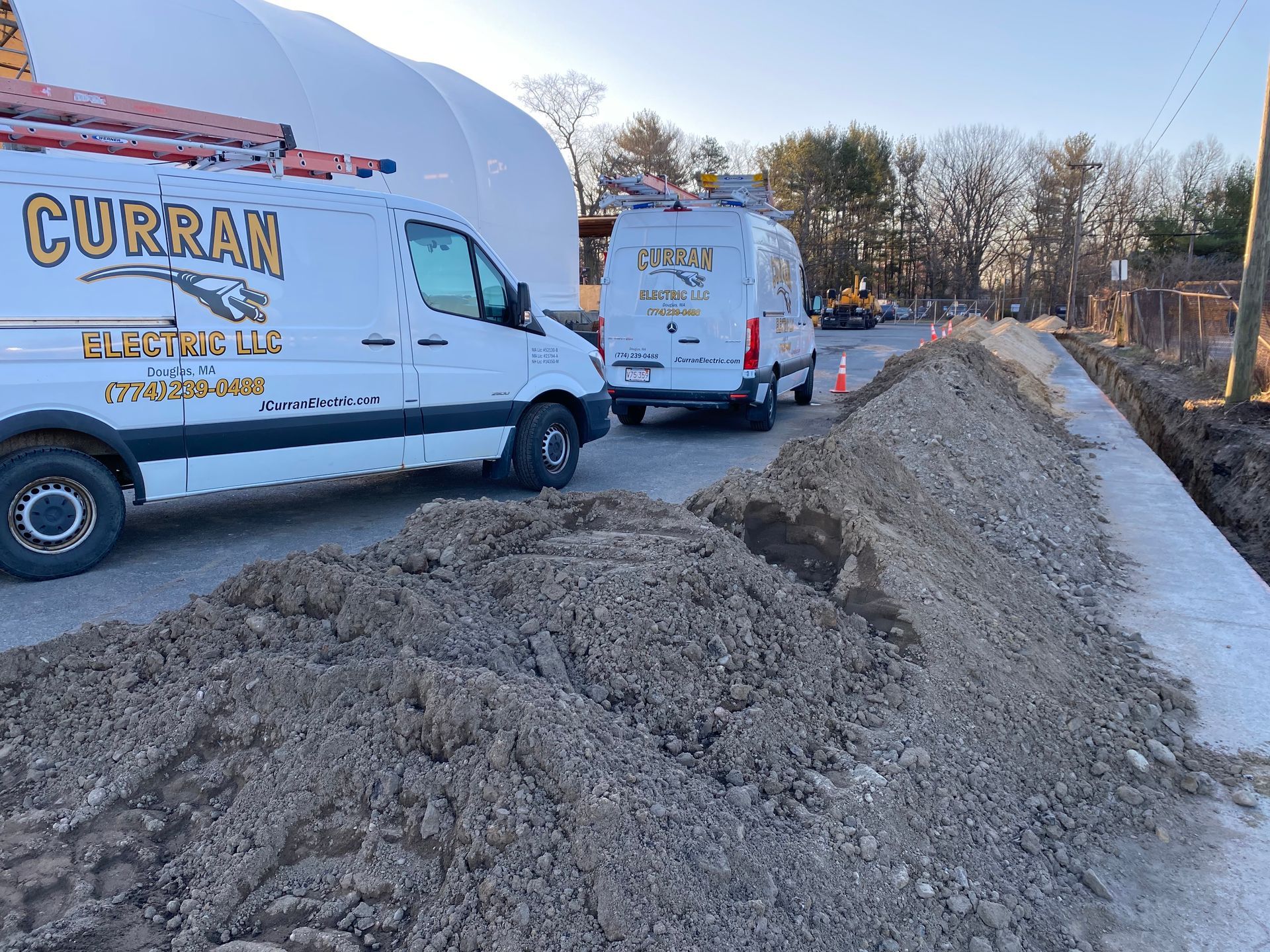 Two white Curran service vans parked next to a pile of dirt on a road next to a white structure and a trench.