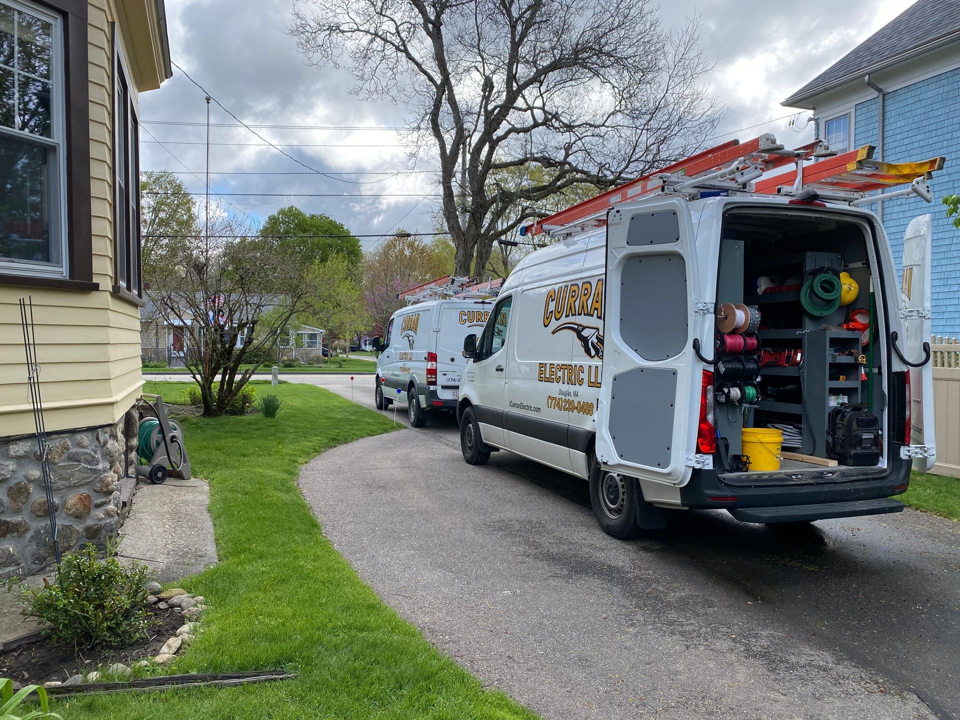 Two service vans parked on a driveway near a yellow house. One van's rear doors are open, revealing tools.
