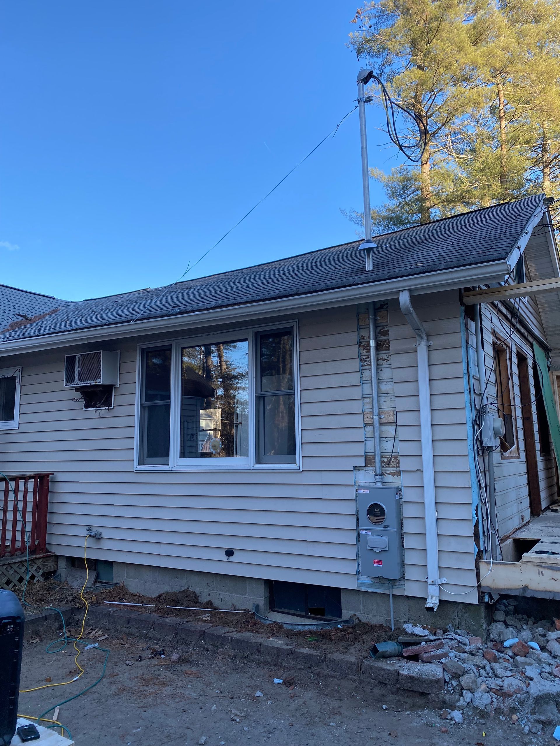 Side view of a house needing repair; light siding, dark roof, electrical box on wall, blue sky.