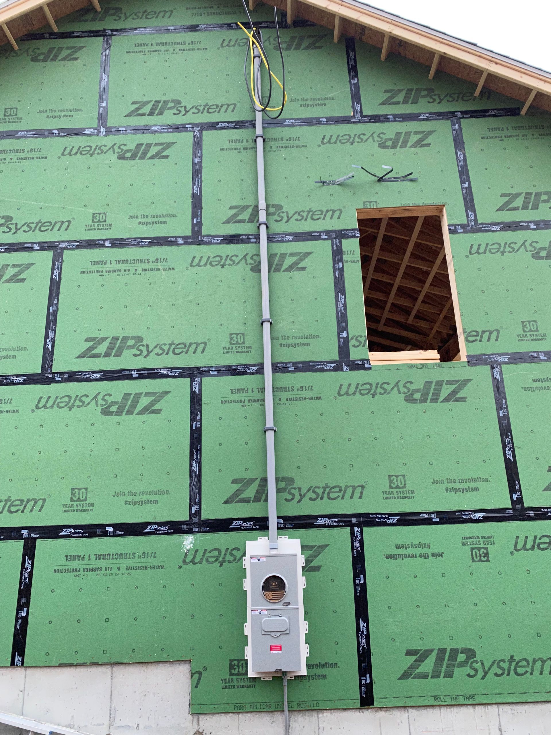 Electrical meter box on a green-sheathed wall with conduit running up to the roof; unfinished construction.