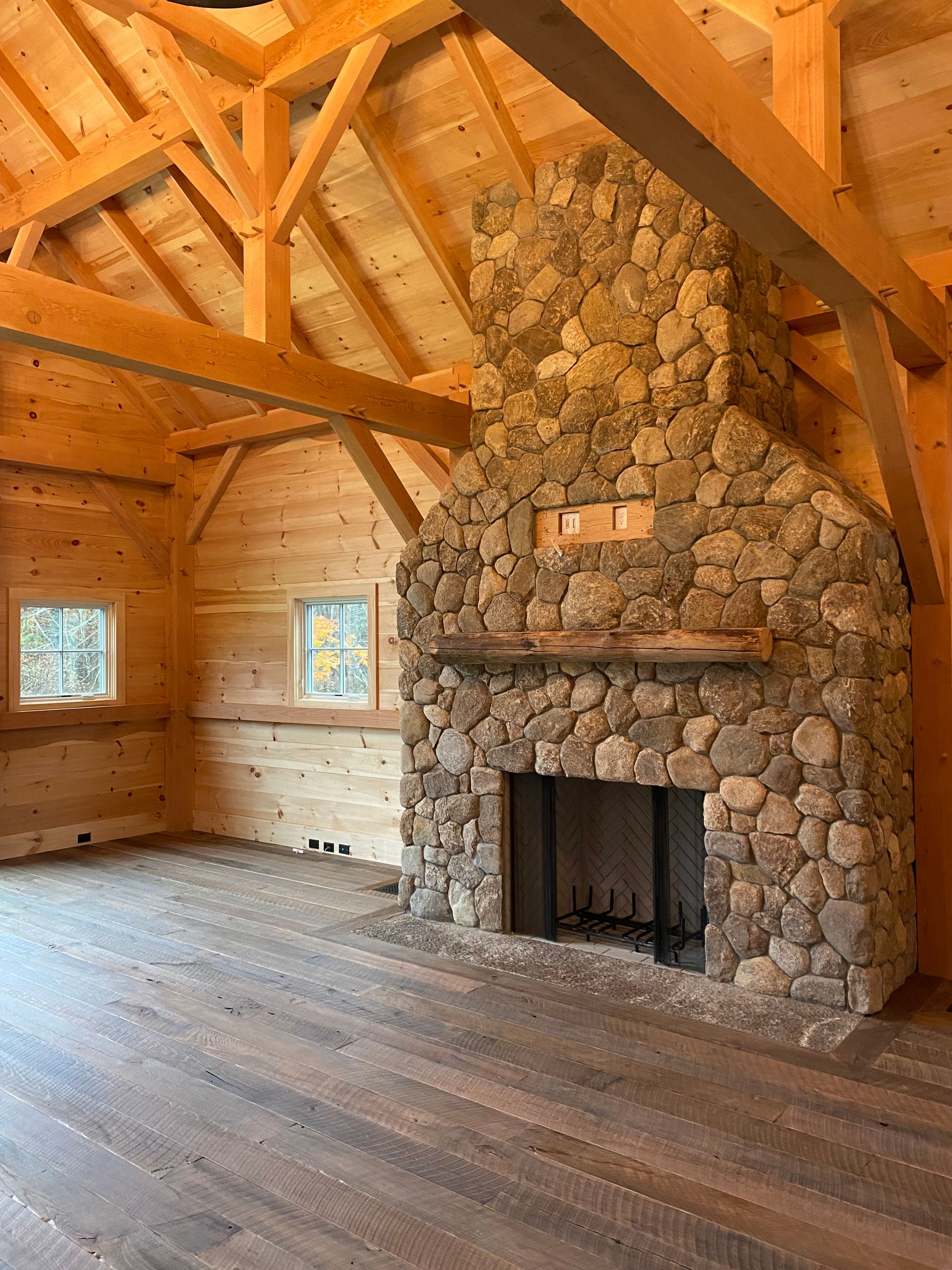 Interior of a rustic room with stone fireplace, wooden beams, hardwood floor, and small windows.