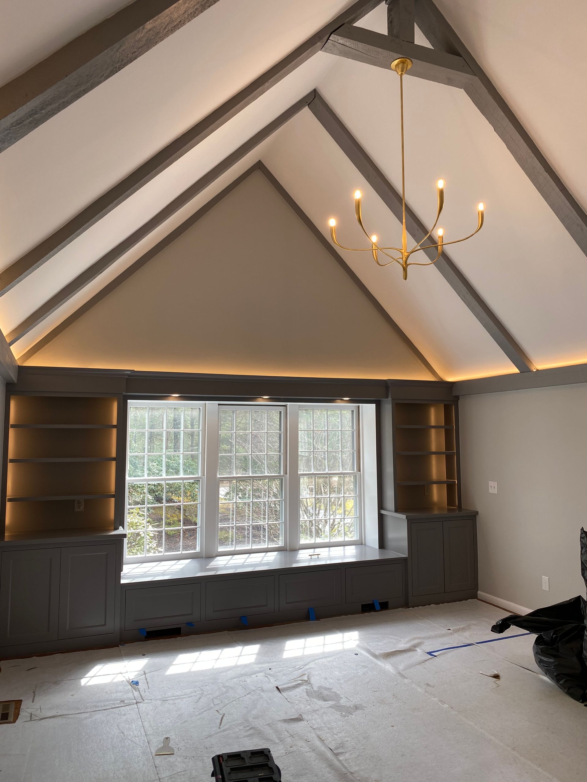Vaulted ceiling with gray beams, gold chandelier, built-in cabinets framing a window with outdoor view, room under construction.