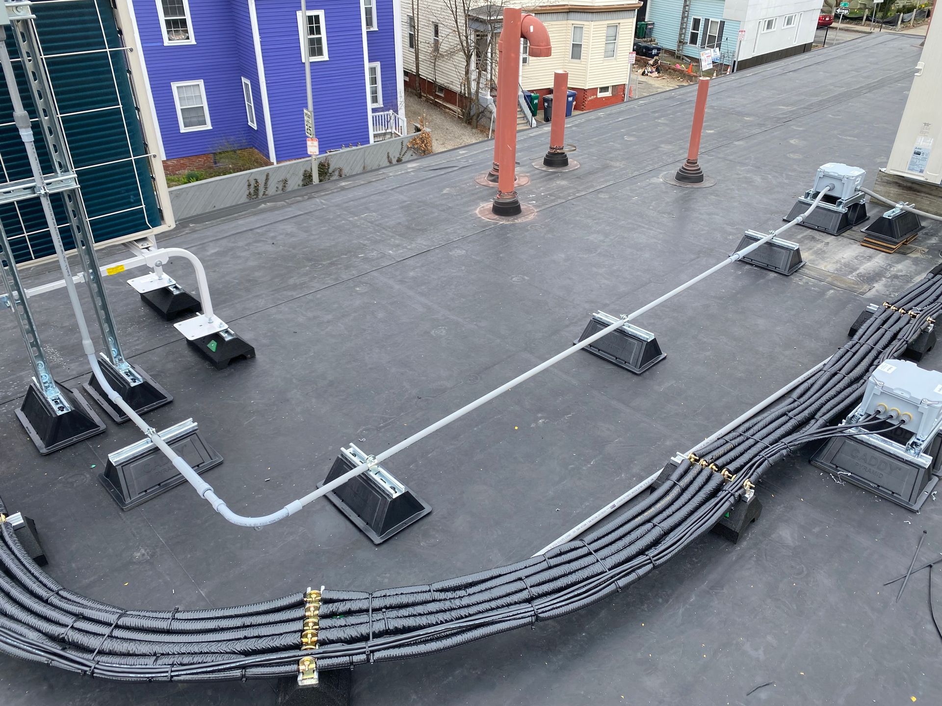 Rooftop with black cable conduits, supports, and vents. Blue and yellow buildings in the background.