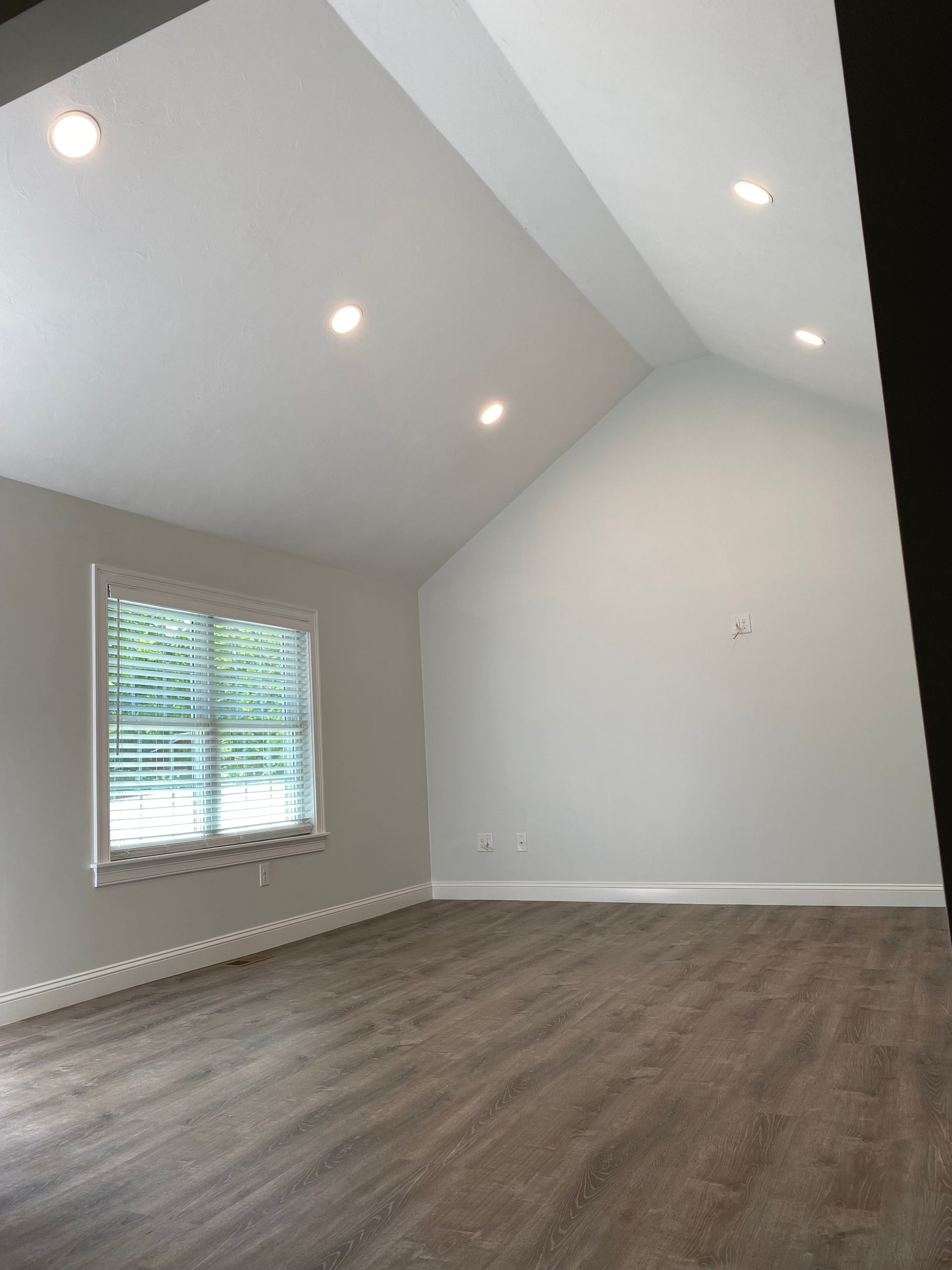 Empty room with vaulted ceiling, recessed lights, window with blinds, and wood-look flooring.