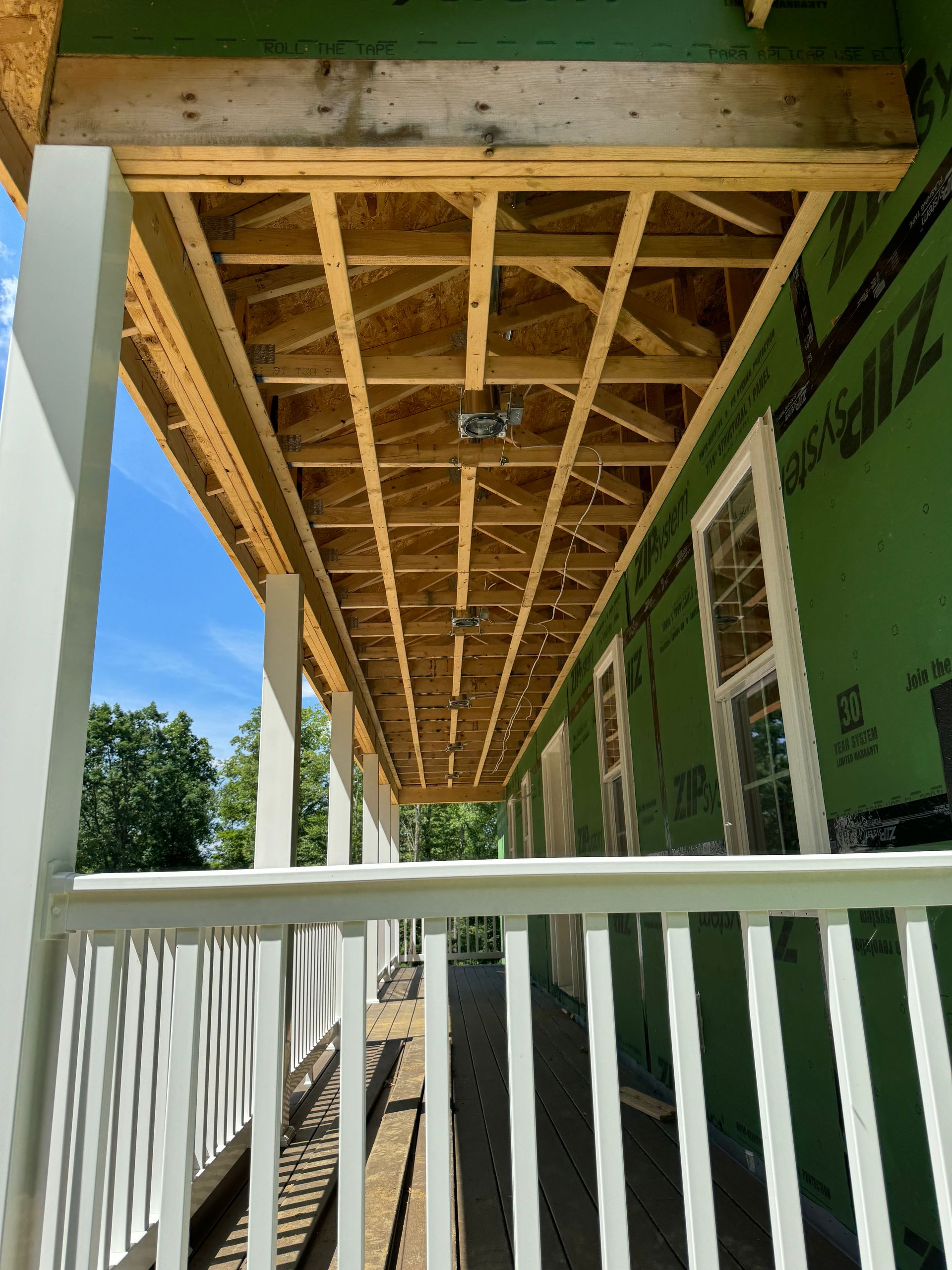 Under-construction porch ceiling with exposed wood framing, light fixture, and white railings. Green building wrap visible.