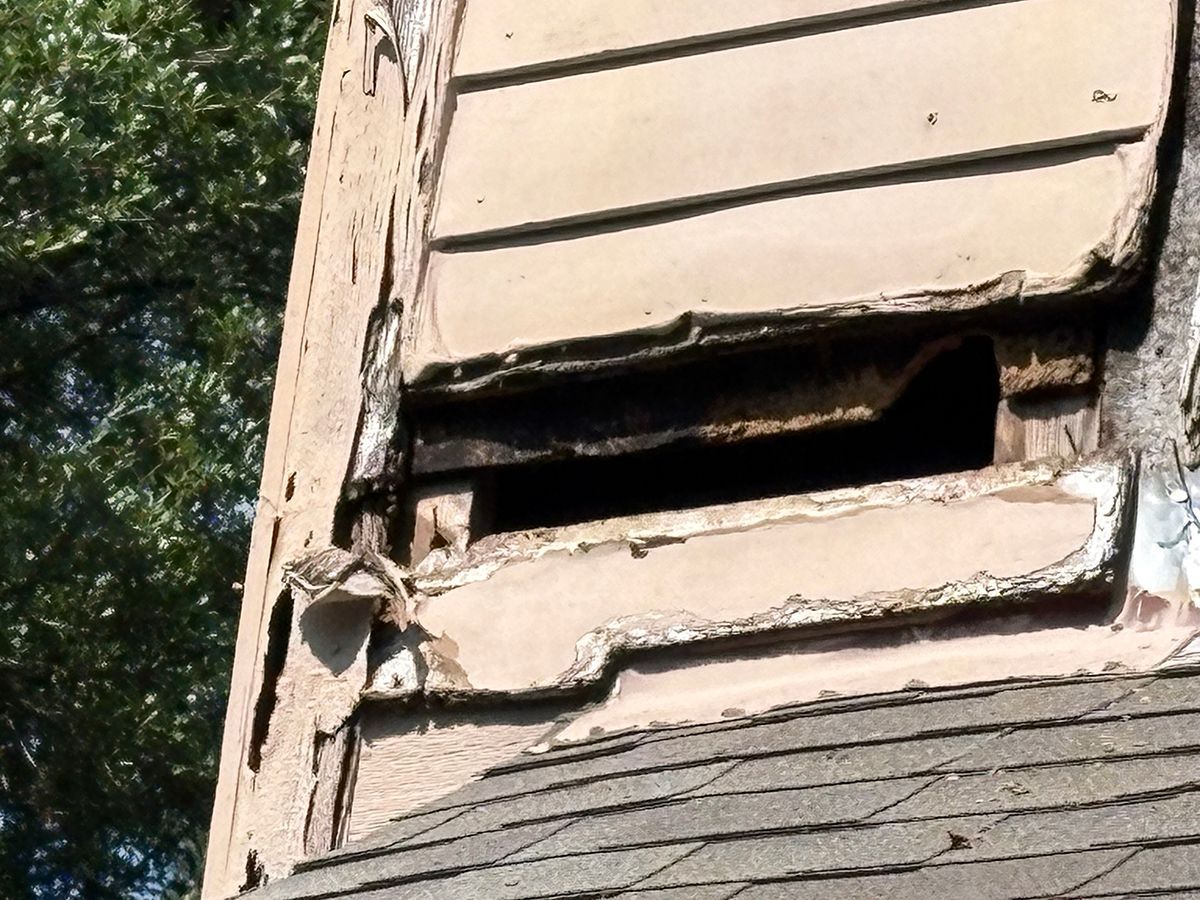 Damaged beige building wall with a broken, open vent and exposed siding near a roofline