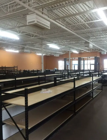 Rows of empty black metal shelving units with light-colored wooden shelves in a large, brightly lit commercial space.