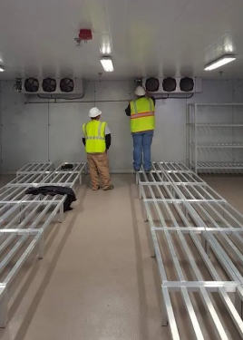 Two technicians in safety vests and hard hats work on cooling units mounted on the wall of a commercial walk-in freezer.