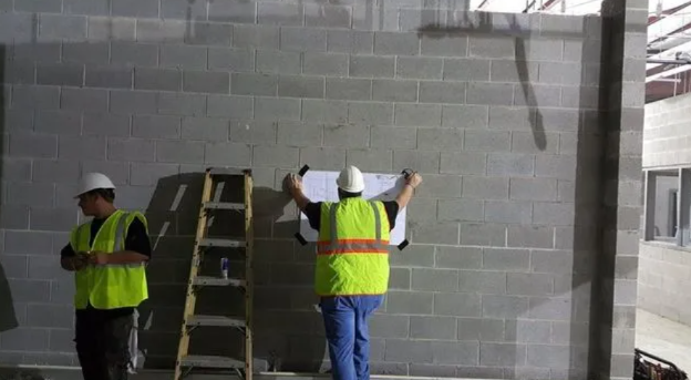 Two construction workers in safety vests and hard hats look at plans posted on a gray concrete block wall.
