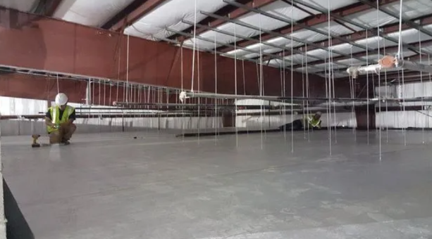 A construction worker wearing a high-visibility vest kneels on a large, level concrete floor inside a steel-framed building.