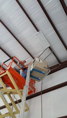 A person in a scissor lift installs a HVAC unit near the ceiling of an industrial building.