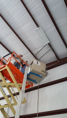 Two workers on an orange scissor lift install a HVAC unit on a white metal ceiling.