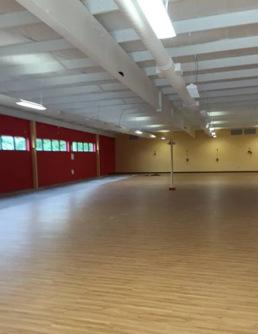 An empty multipurpose room with light wood floors, high white-beamed ceilings, and one wall painted red with windows.