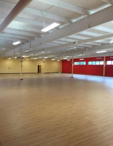 A large, empty hall with light wood flooring, white ceiling beams, fluorescent lights, and one bright red wall.