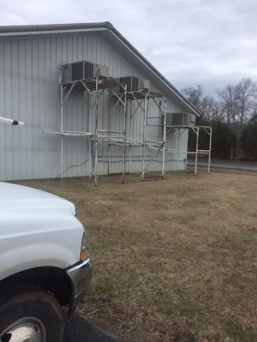 Three outdoor air conditioning units mounted on metal support structures against the side of a grey metal building.