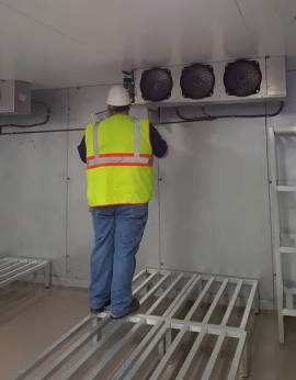A worker in a high-visibility vest stands on metal shelving to inspect a ceiling-mounted industrial cooling unit.