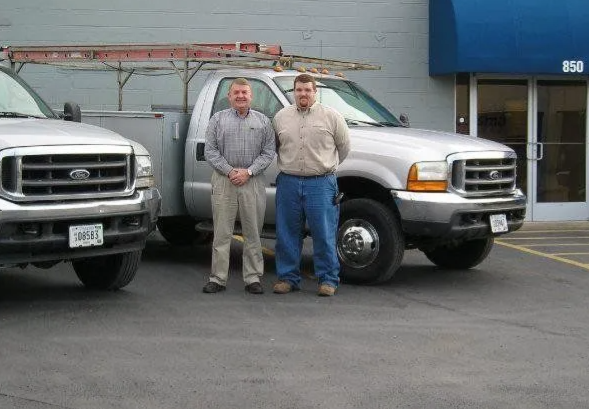 Two men stand in front of two silver utility trucks parked outside a building with a blue awning.