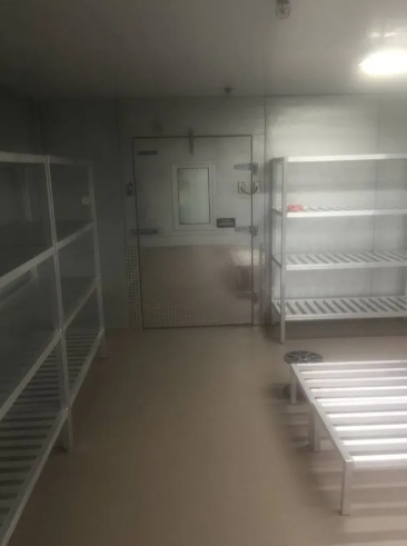 An empty walk-in cooler or storage room featuring white metal shelving units and a single stainless steel door.