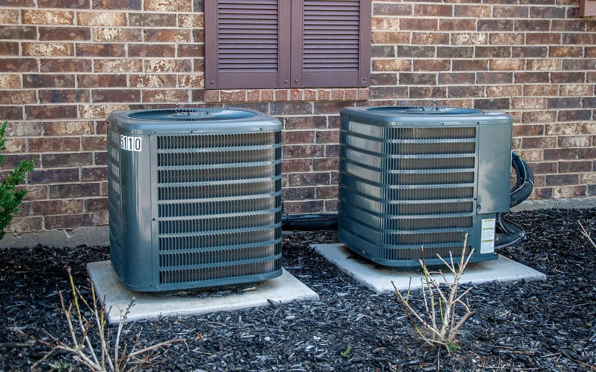 Two grey outdoor air conditioning units sit side-by-side on concrete pads against a brick wall.