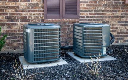 Two grey outdoor air conditioning units sit side-by-side on concrete pads against a brick wall.