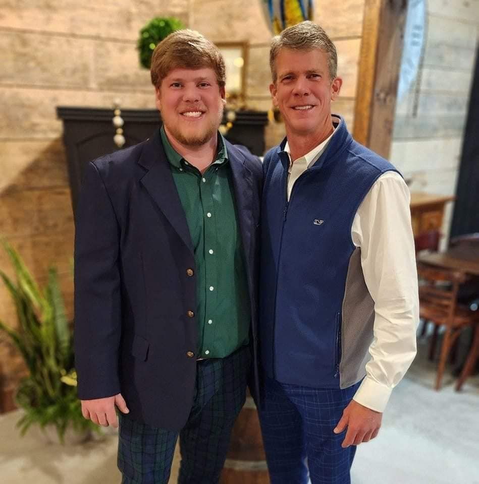 Two men stand indoors, one in a blazer and green shirt, the other in a vest and white shirt.