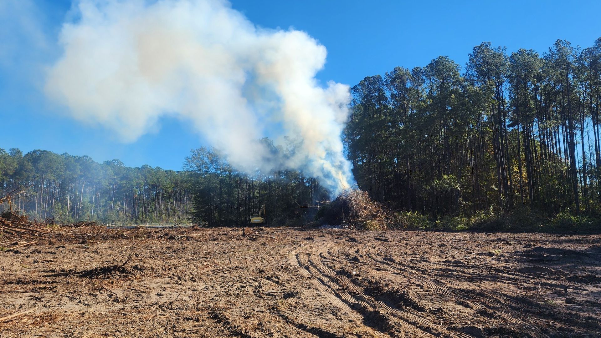 Smoke billows from a burning pile of debris next to a treeline; clear blue sky, brown earth.