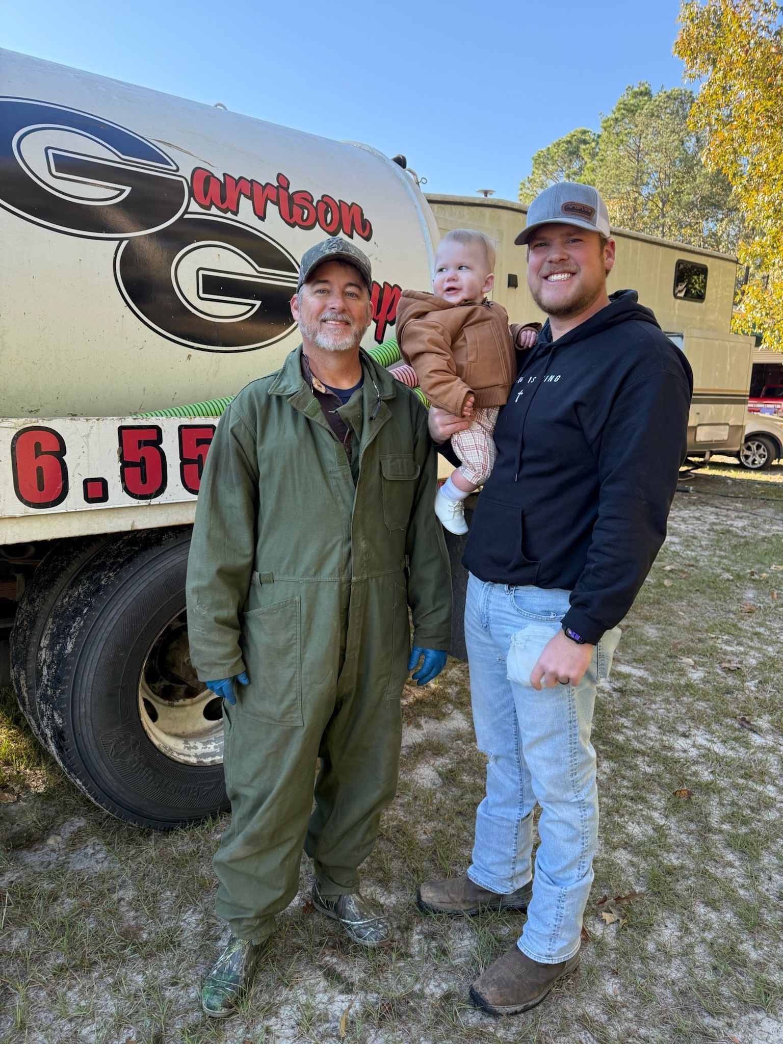 Three people stand by a truck with a logo. One in green jumpsuit, one holding a baby, and one in a black hoodie. Outdoor setting.