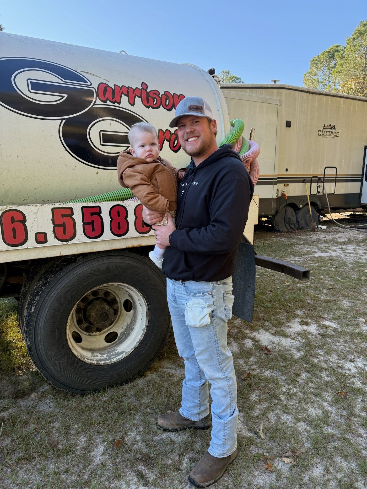 Man holding a baby in front of a water truck. Man wears jeans, boots, hoodie, and a cap. Baby in a brown jacket.