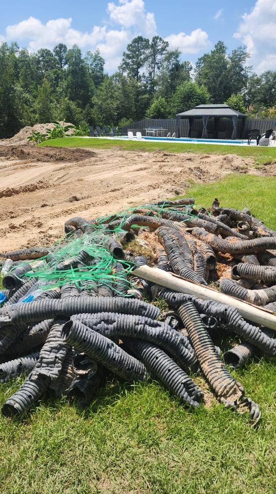 Pile of drainage pipes and netting on grass, dirt patch in foreground. Pool and trees in background.