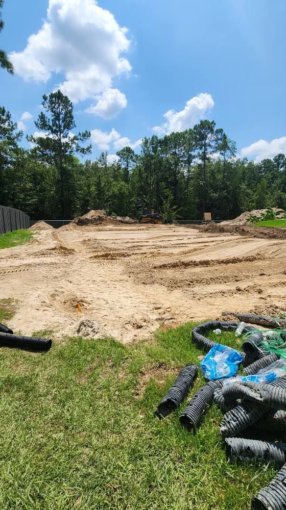 A cleared dirt lot in a yard, trees in the background, blue sky with clouds, drainage pipes in foreground.