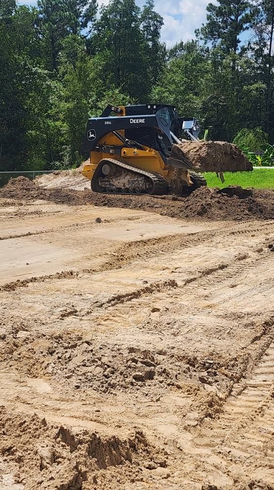 John Deere skid steer moving dirt on a construction site; brown earth, green trees in background.