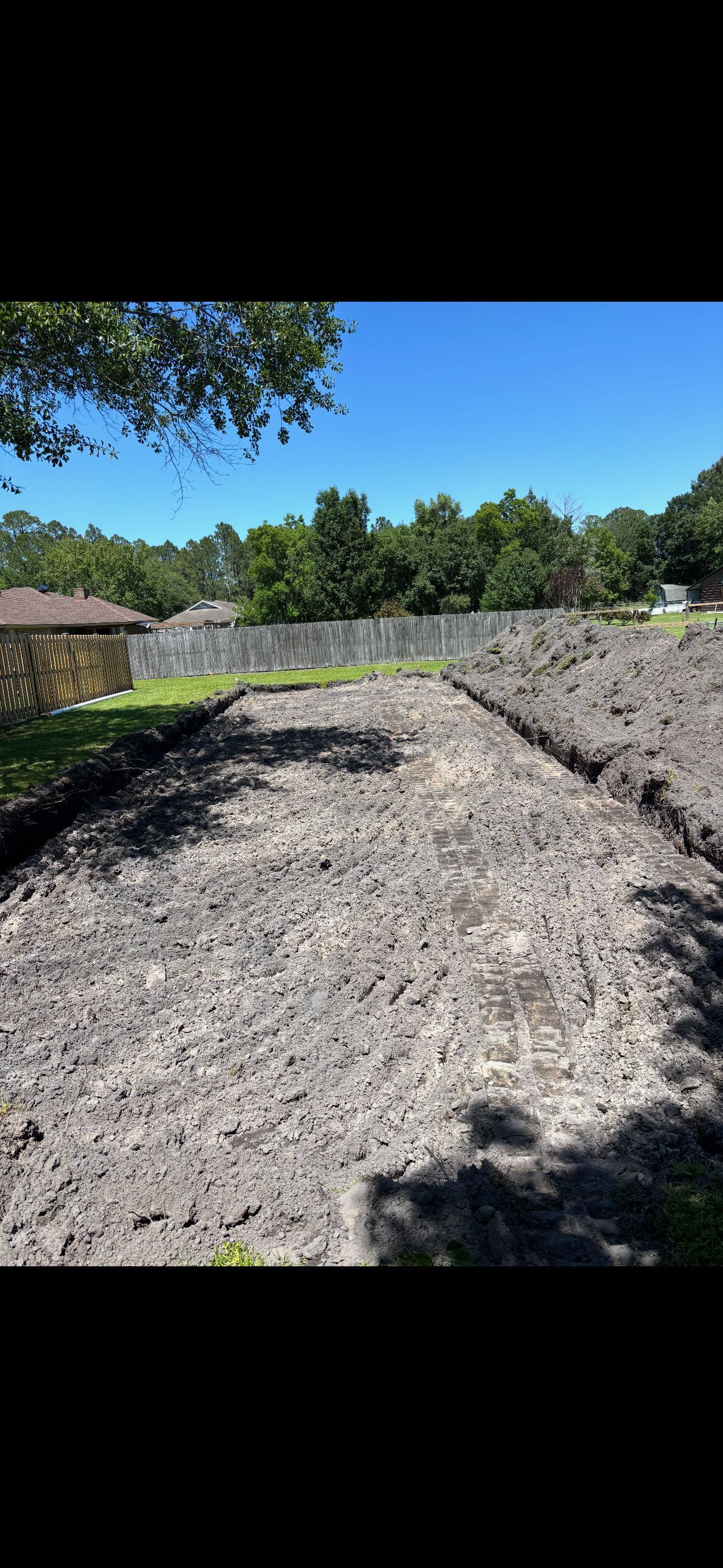 A backyard featuring a long, rectangular plot of freshly tilled, dark soil under a bright blue sky and sunny conditions.