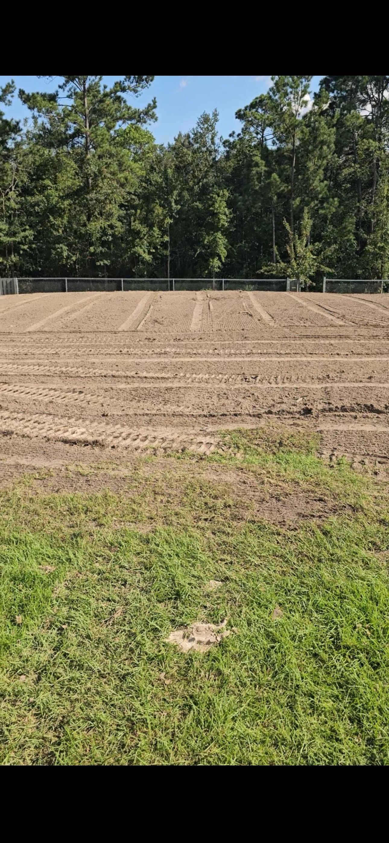 A freshly tilled, empty field with visible planting rows, bordered by a green lawn and a line of trees in the background.