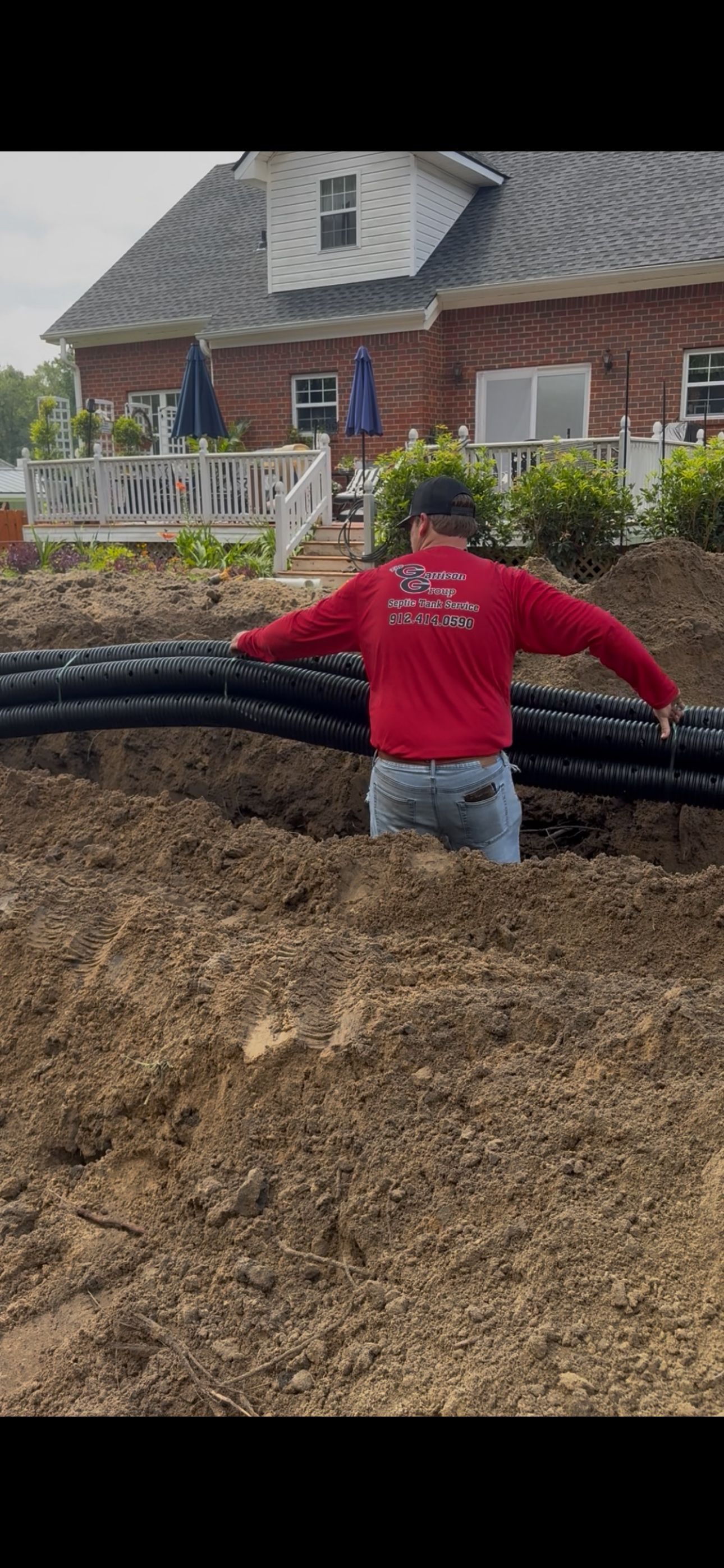 A person in a red shirt stands in a muddy trench, installing long black corrugated pipes near a brick house.