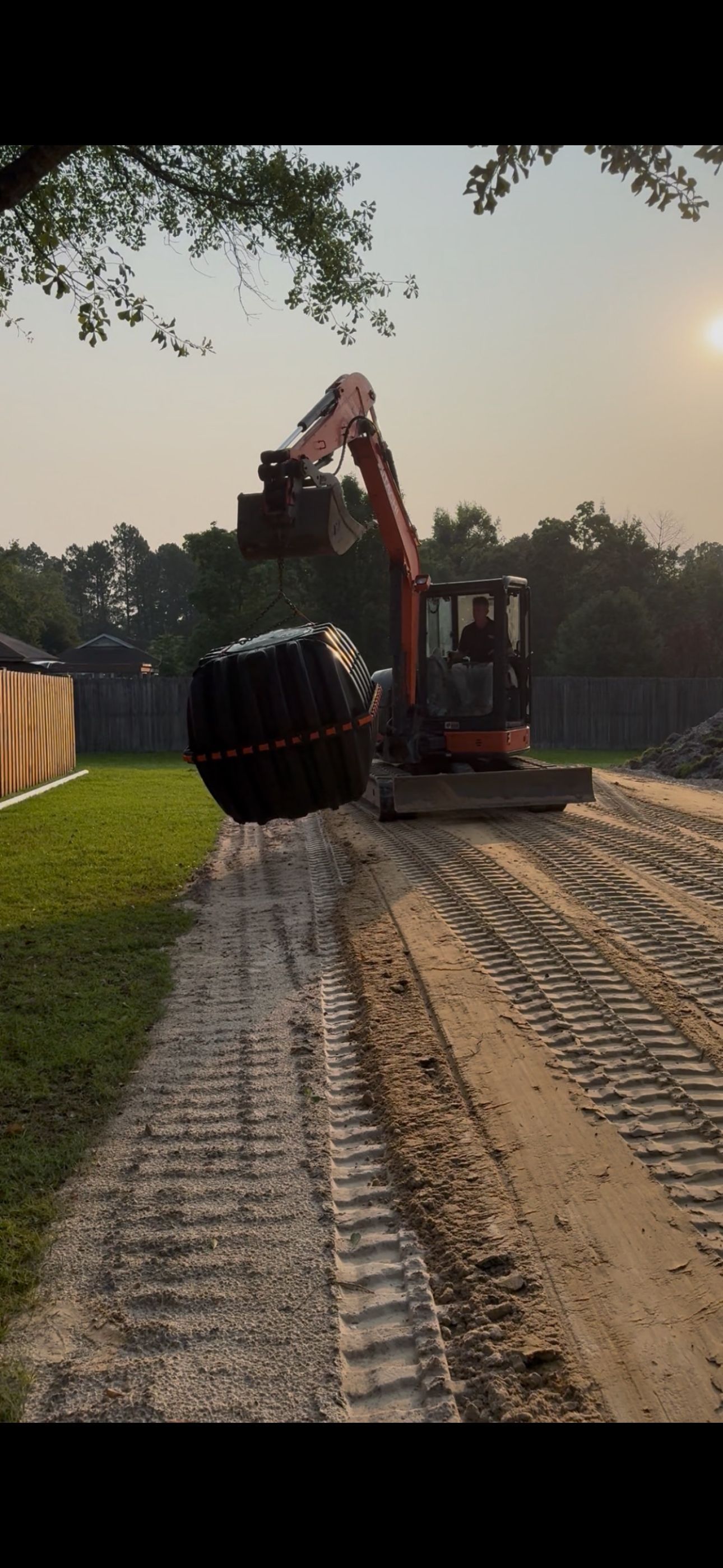 An orange excavator lifts a large, strapped plastic culvert pipe on a dirt construction site.