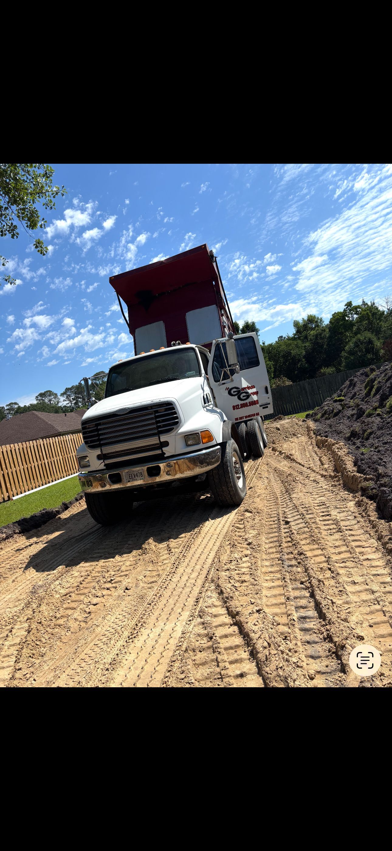 A white dump truck with its bed raised, parked on a dirt path at a construction site under a clear blue sky.