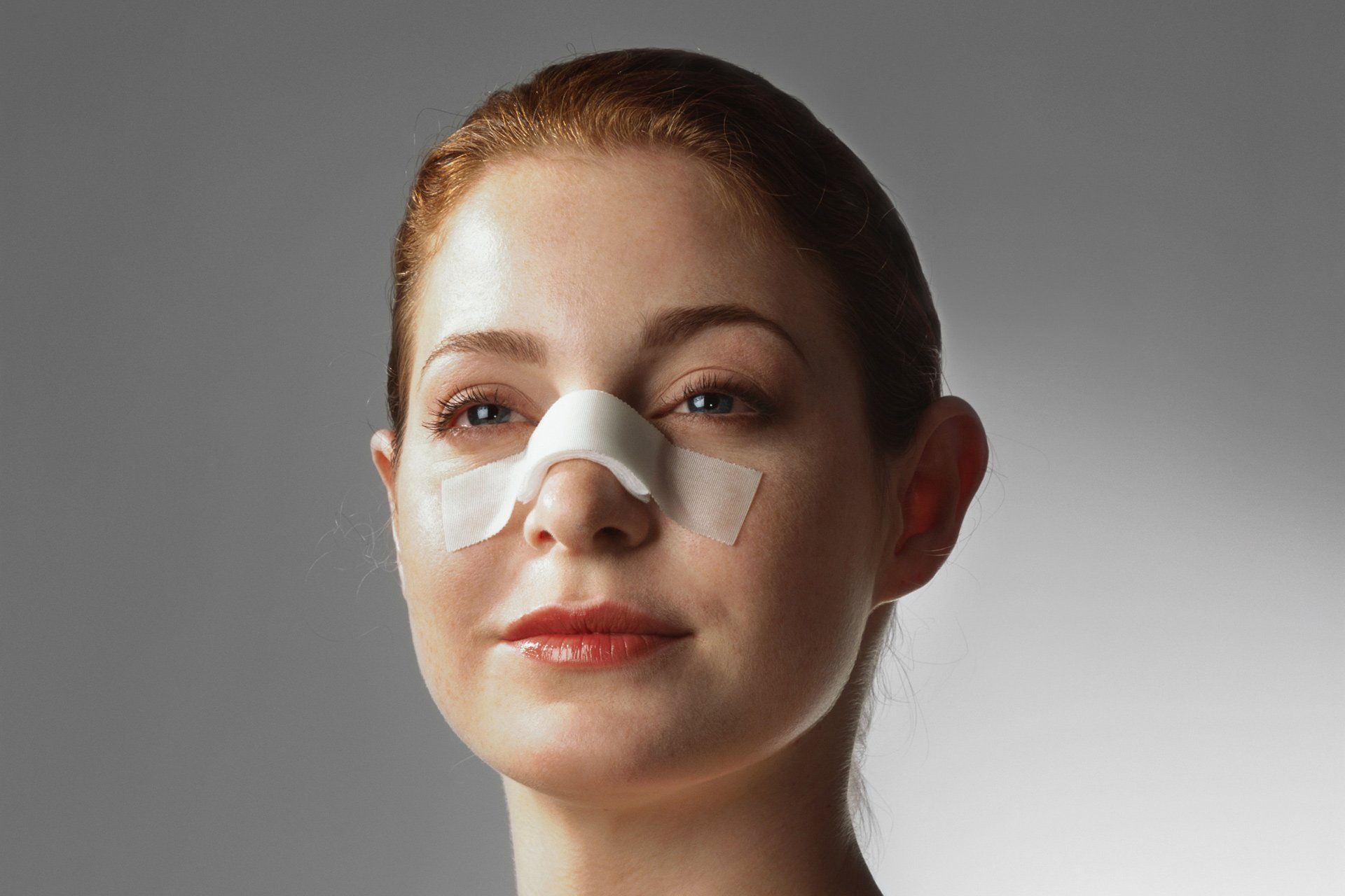 A woman with a bandage on her nose is looking at the camera.