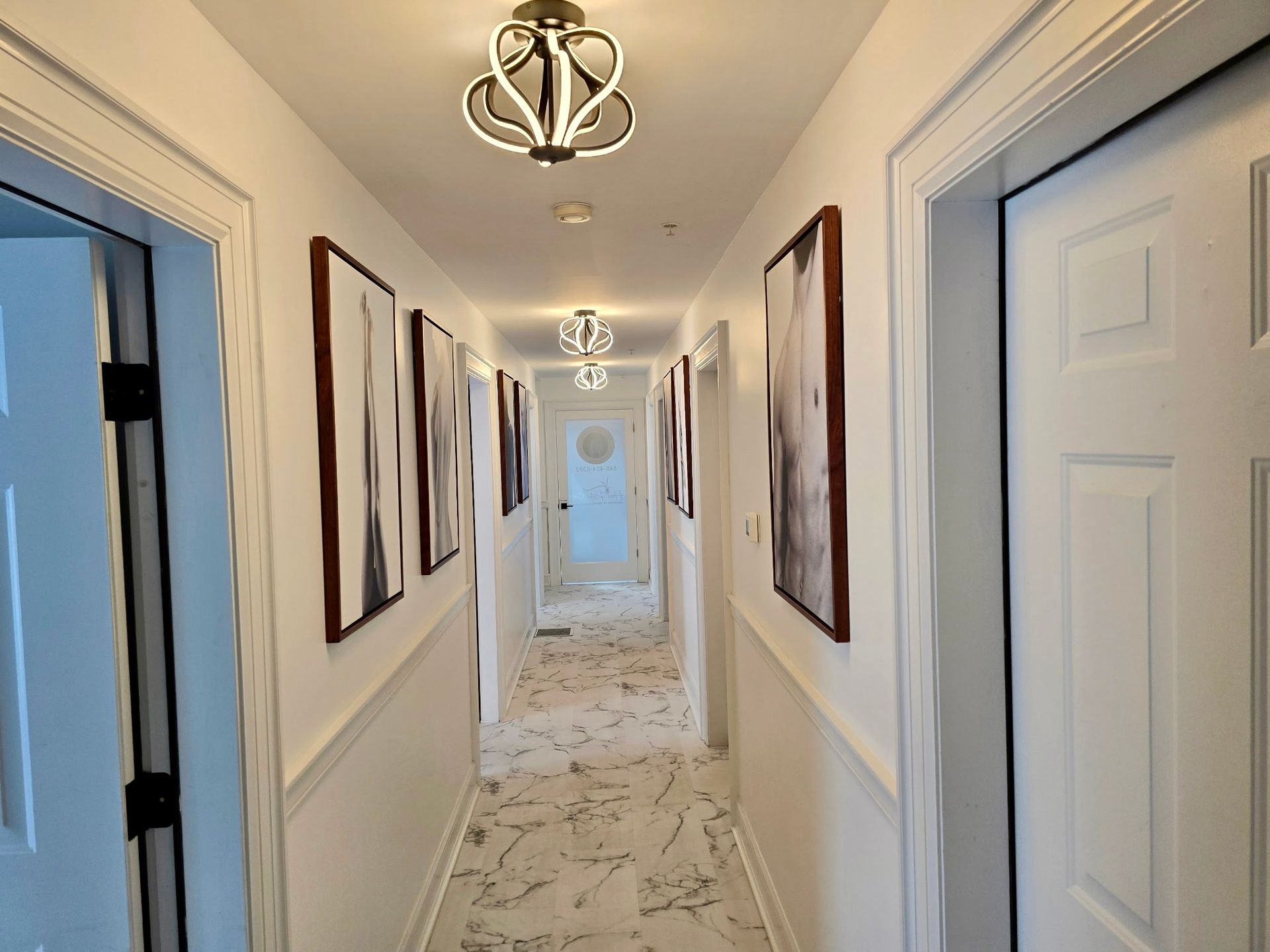 Narrow white hallway with artwork, ornate lighting, and patterned floor.