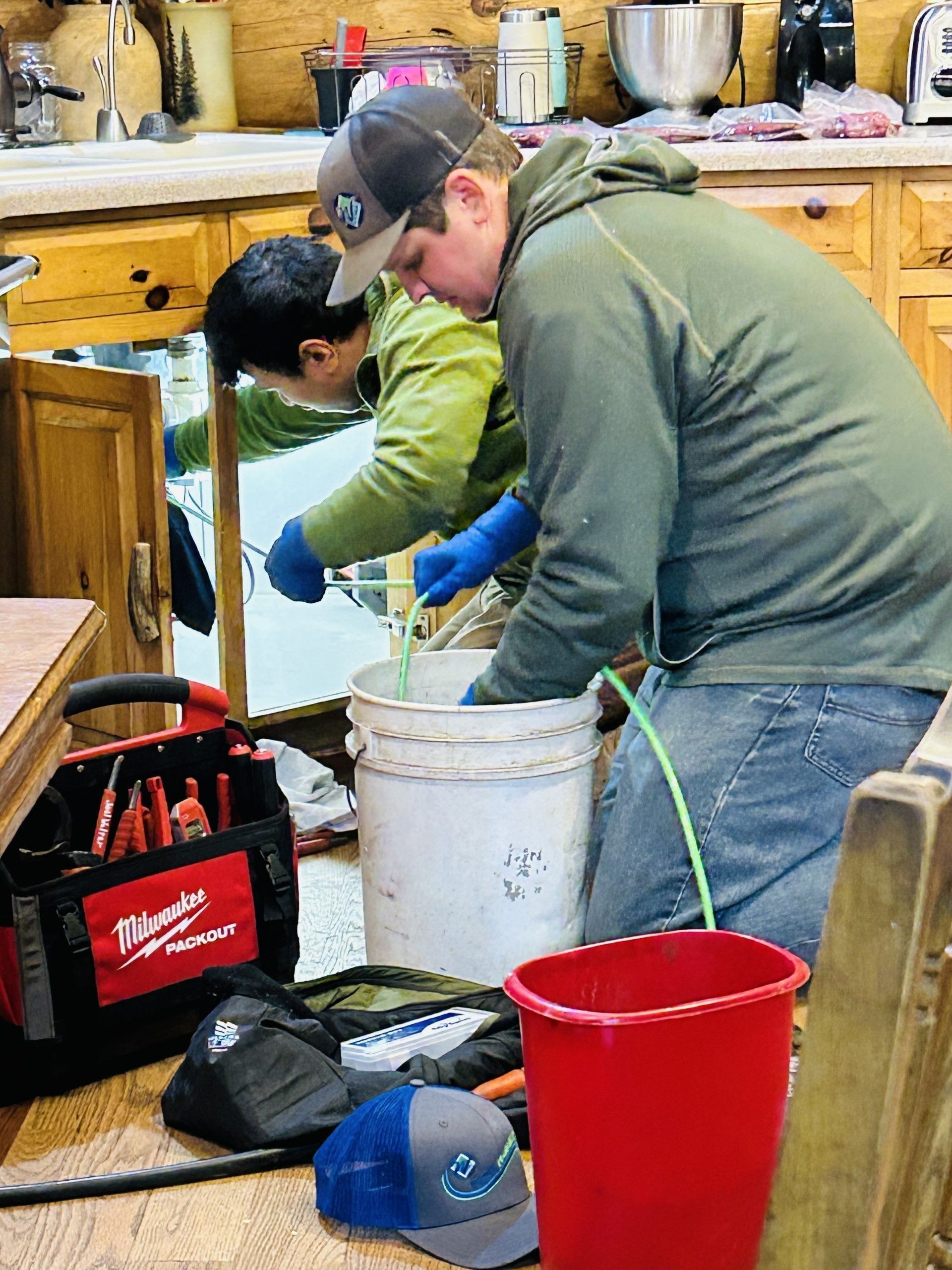 Two men are performing maintenance on the kitchen sewage system.