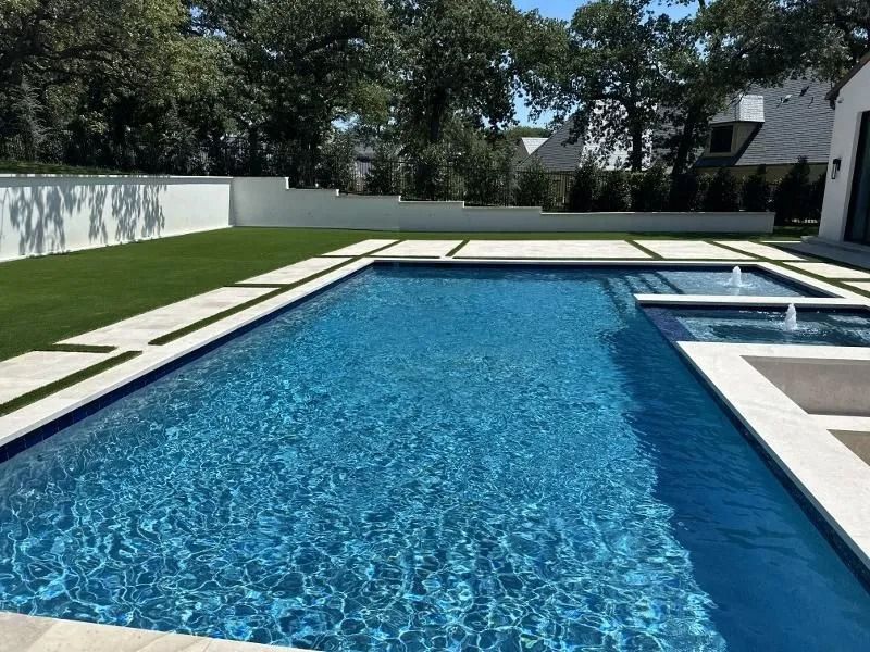 Rectangular blue pool with white trim and a small spa. Artificial turf and a white wall in background.