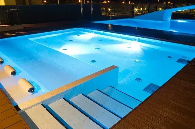 A rectangular, blue-lit pool with integrated steps and jets. Another pool is visible in the background, also with blue lighting.